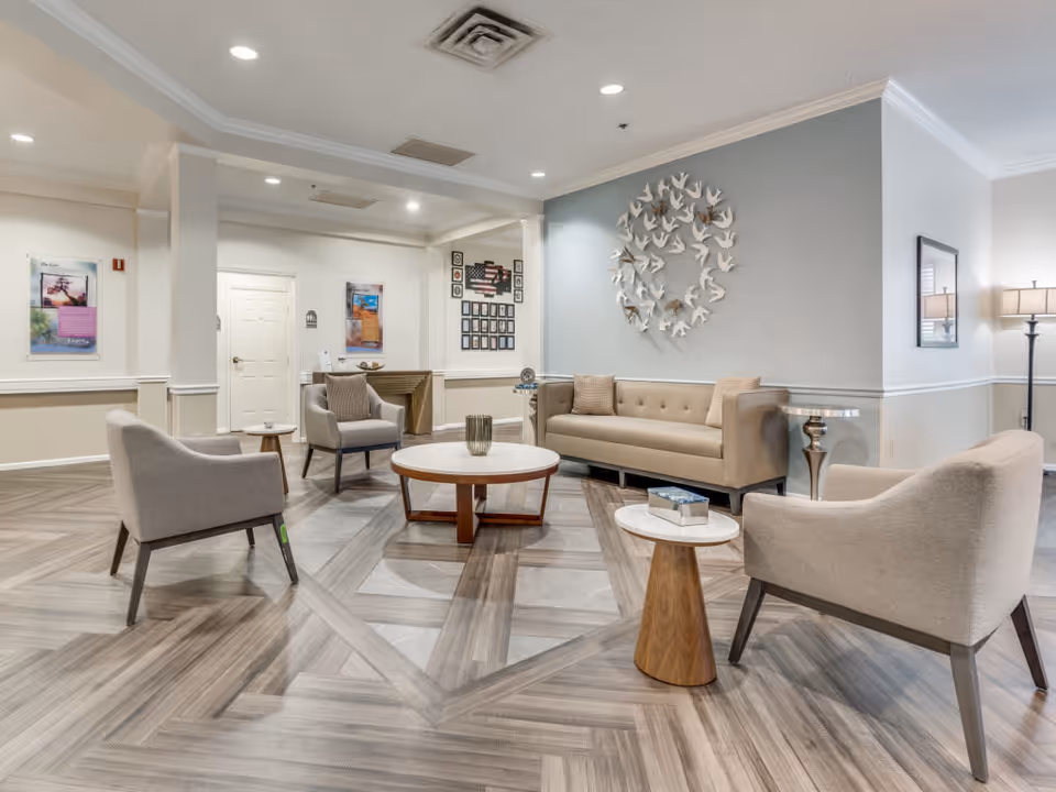 A modern and spacious living room area in a senior living facility featuring a beige sofa, three beige armchairs, a round wooden coffee table, and two small side tables. The room has light-colored walls with decorative wall art, recessed ceiling lights, and a floor lamp in the corner. The flooring is a patterned wood design.