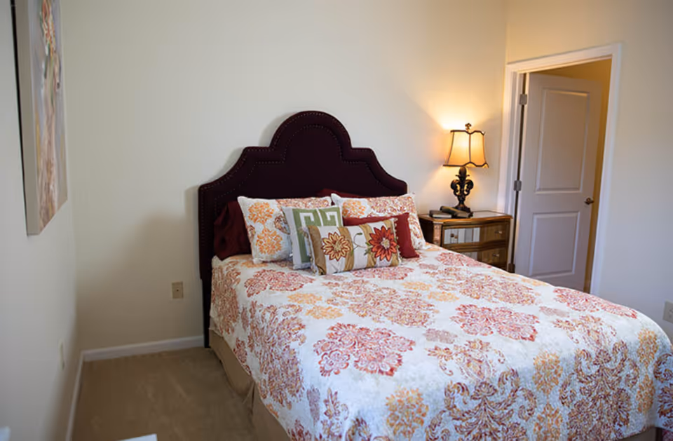 A bedroom with a bed featuring a dark purple upholstered headboard and a floral patterned bedspread in shades of orange, red, and beige. There are multiple decorative pillows on the bed. A wooden nightstand with a lamp and a phone is next to the bed. The walls are light-colored, and there is a door open to another room.