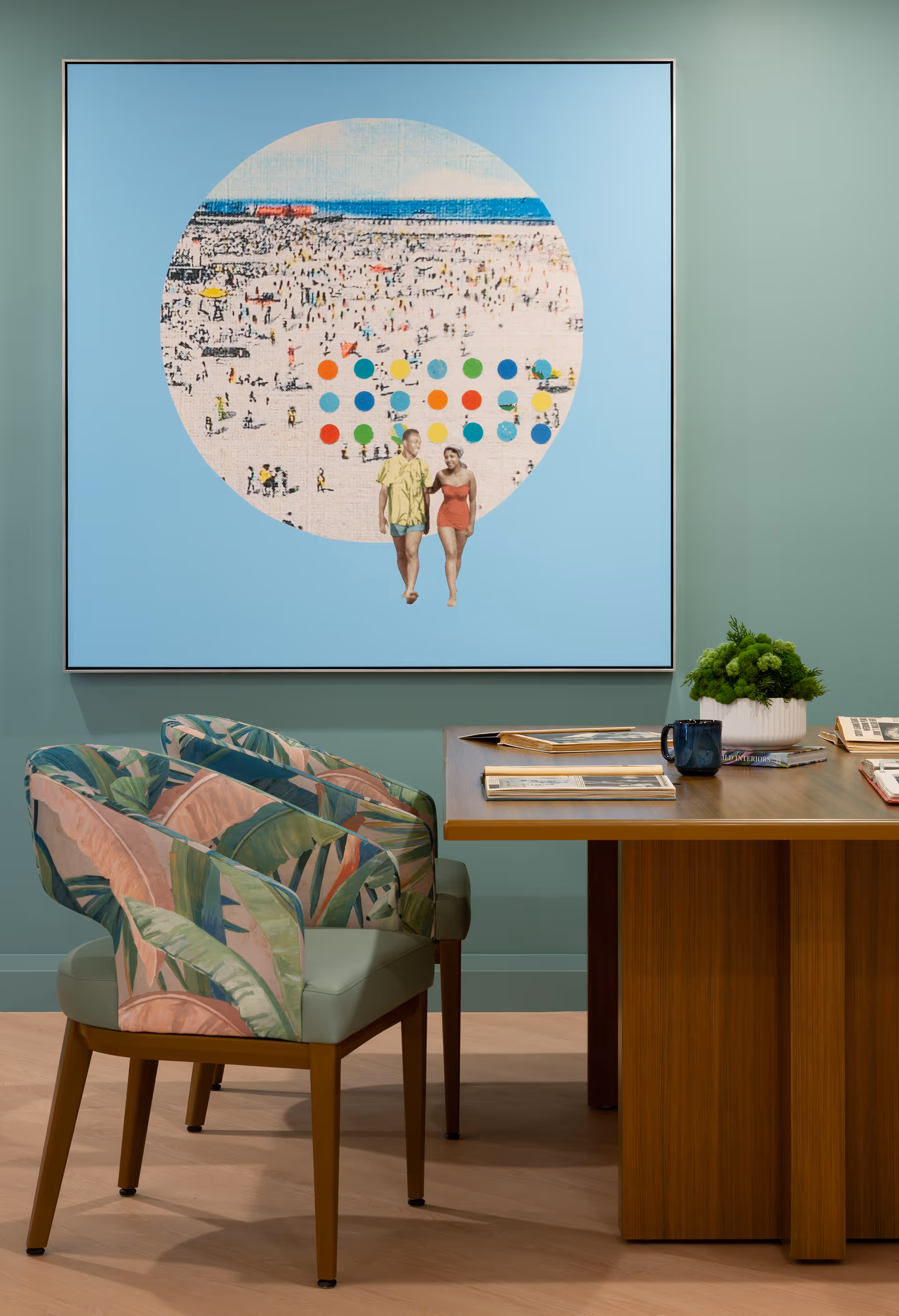 A modern interior room with a wooden table and two chairs featuring tropical leaf-patterned upholstery. On the wall above the table is a large framed artwork depicting a beach scene with many people and two individuals walking in the foreground. The wall is painted in a soft green color, and there is a small green plant on the table along with some books and a blue mug.