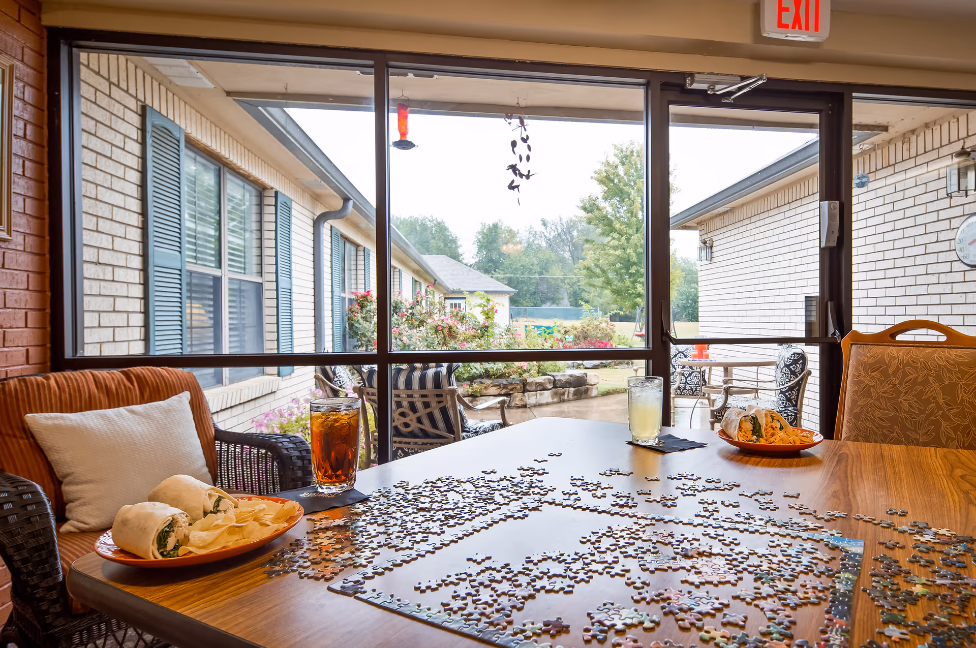 A wooden table with a partially completed jigsaw puzzle and two plates of wraps with potato chips. Two drinks, one iced tea and one lemonade, are placed on black napkins. The table is surrounded by cushioned chairs and is positioned near large glass sliding doors that open to an outdoor patio with garden and seating. The scene is bright with natural light coming through the glass doors.
