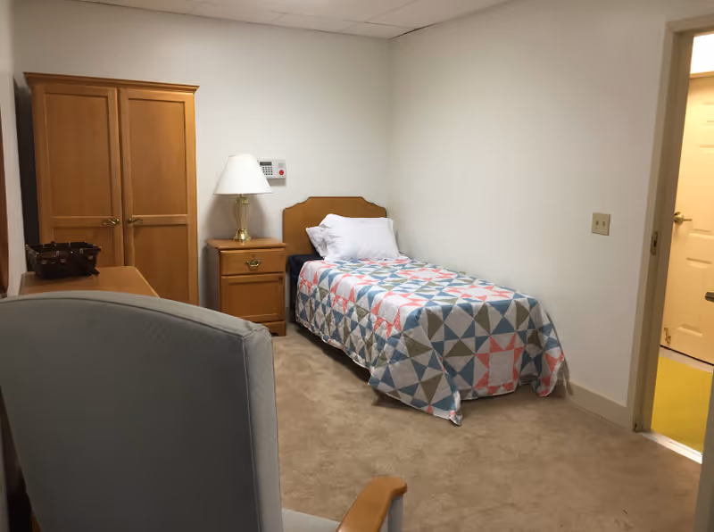 A small bedroom with a single bed covered with a colorful geometric patterned quilt. Next to the bed is a wooden nightstand with a lamp on it. There is a wooden wardrobe and a desk with a basket on top. A cushioned chair with wooden armrests is partially visible in the foreground. The walls are plain white and the floor is carpeted. A door is open, showing a glimpse of a yellow floor in the adjacent room.