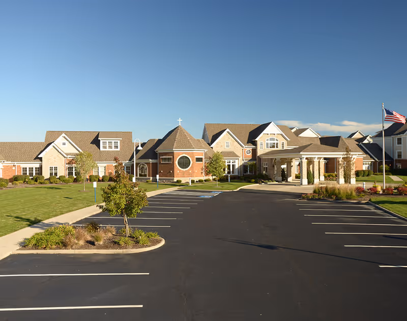 Front exterior of a senior living facility with a large parking lot, landscaped entrance, and an American flag under a clear sky.
