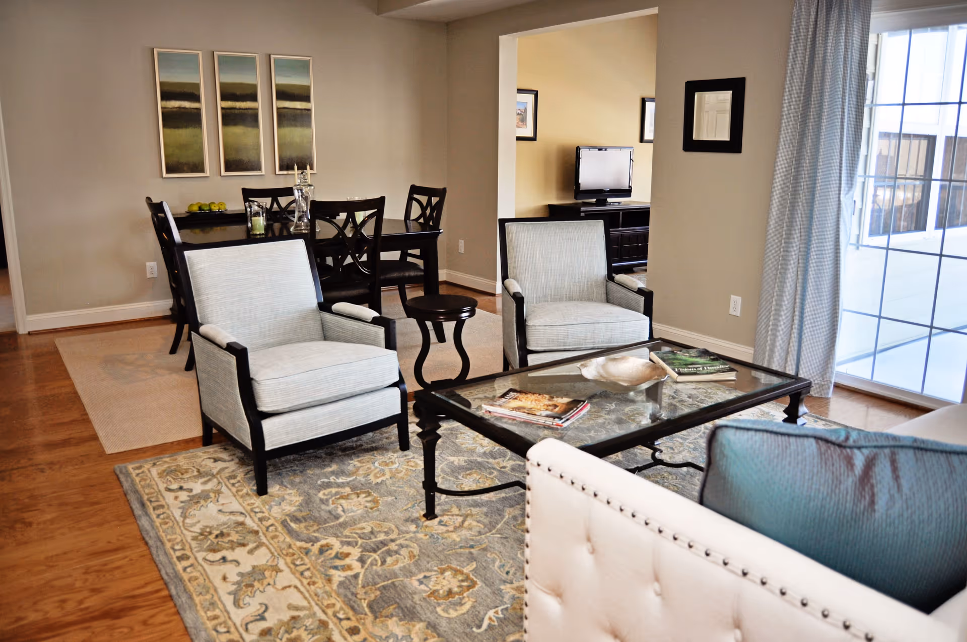 Bright furnished living room with two upholstered armchairs around a glass coffee table on a patterned rug and a dining table visible in the background.