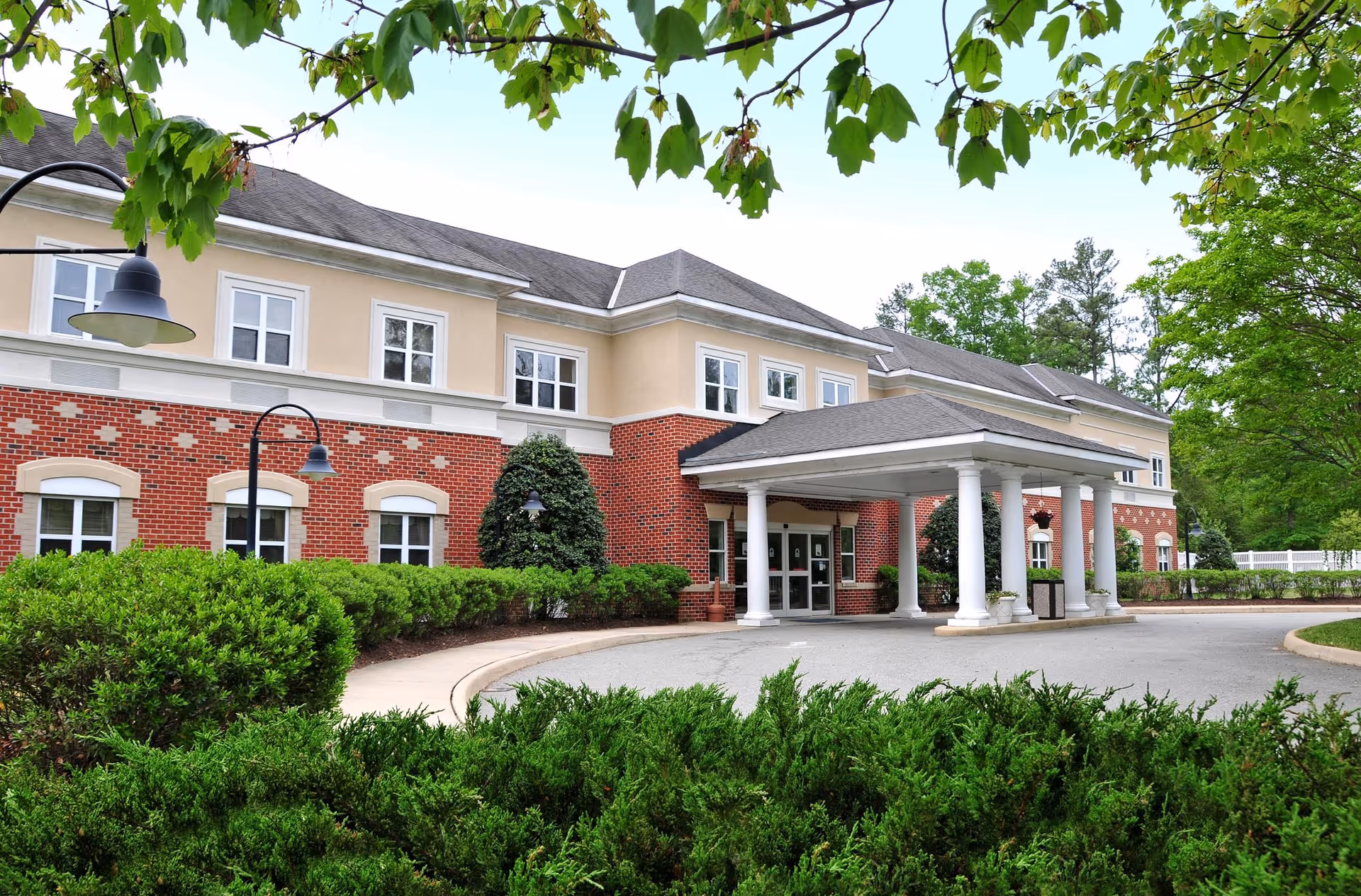 Exterior view of The Crossings at Ironbridge senior living facility showing a two-story building with a combination of red brick and beige siding. The entrance features a covered drop-off area supported by white columns. The surrounding area includes well-maintained green bushes, trees, and a curved driveway.