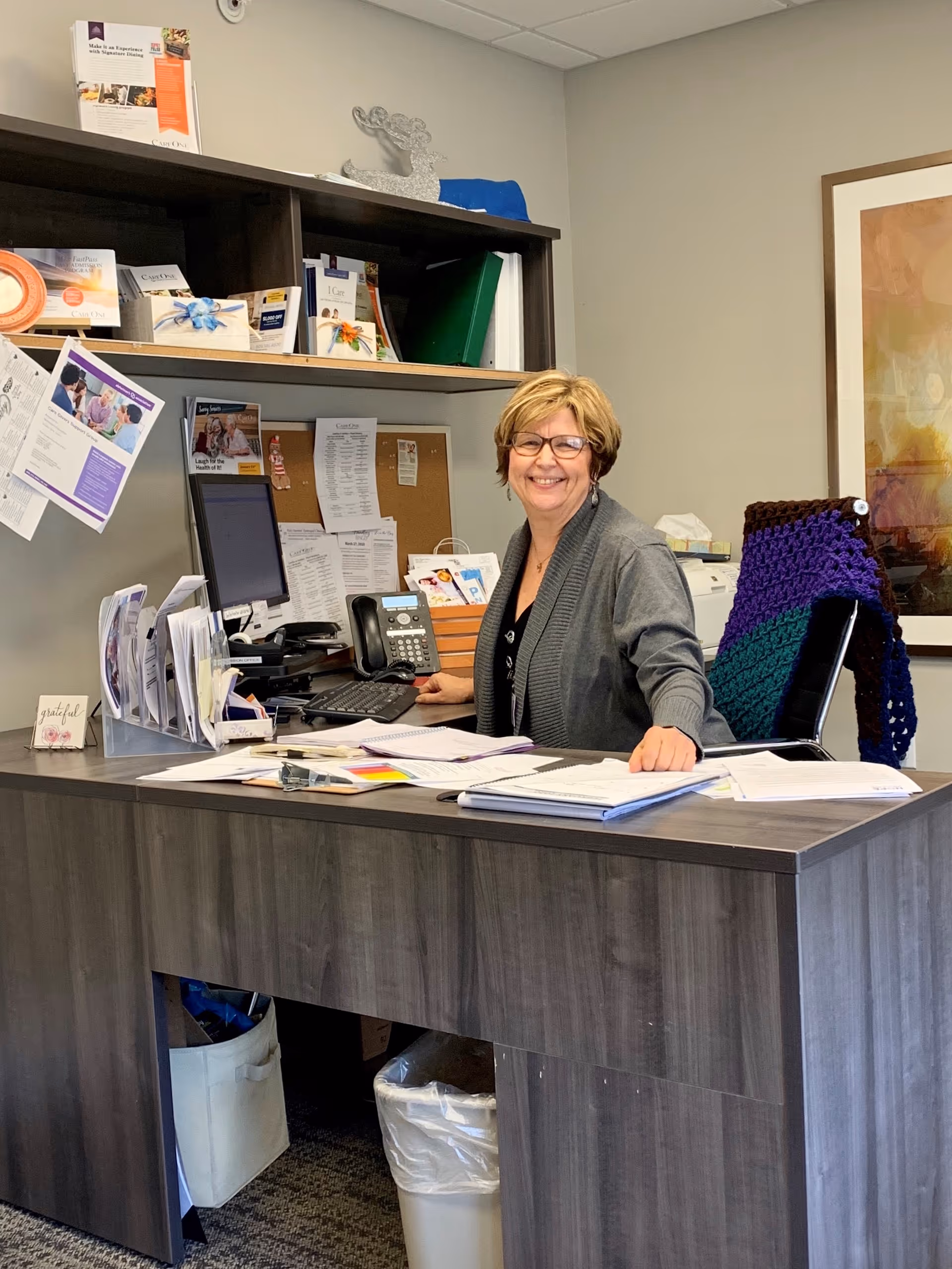 A smiling woman with short blonde hair and glasses sits at a desk in an office. The desk is cluttered with papers, a computer, a phone, and office supplies. Behind her is a bulletin board with pinned papers and shelves holding books and decorative items. A colorful knitted blanket is draped over the back of her chair.