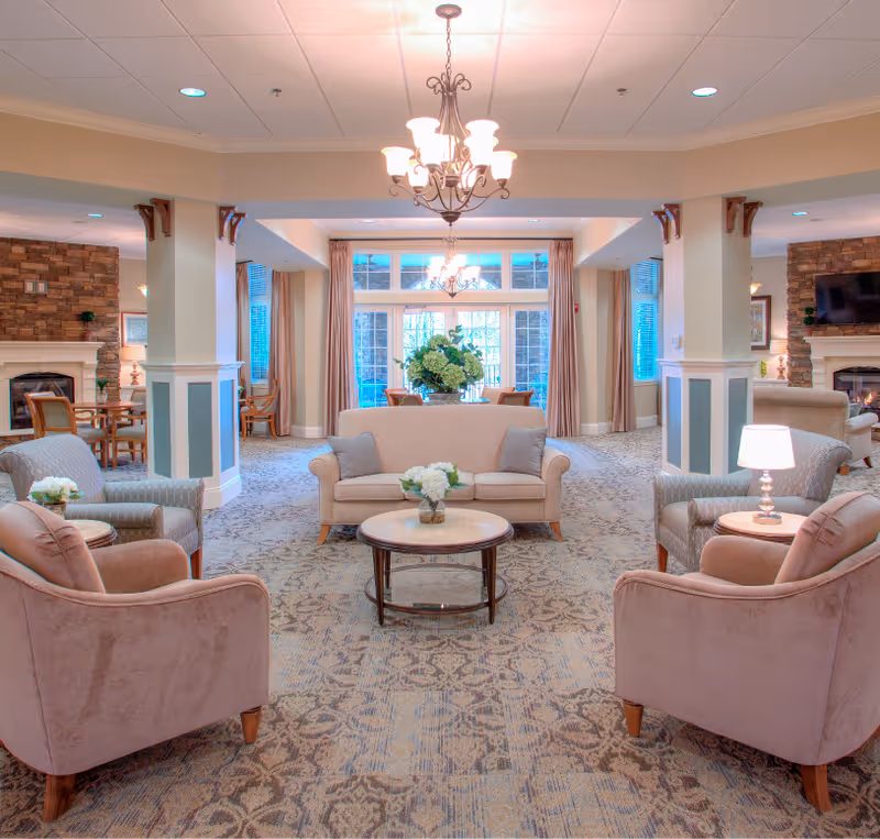Bright, spacious assisted-living common lounge with sofas, armchairs, coffee tables, chandeliers and large windows.