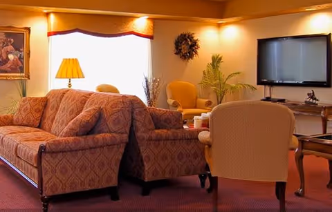 A cozy living room with patterned sofas and armchairs arranged around a coffee table. The room is warmly lit with a table lamp and recessed ceiling lights. A large flat-screen TV is mounted on the wall, and decorative elements like a framed painting, a wreath, and a potted plant add to the inviting atmosphere.