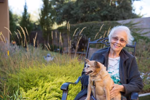 An elderly woman with white hair and glasses sitting on a black metal chair outdoors, smiling and holding a small tan dog on her lap. The background features green shrubs, tall grasses, and trees in a garden setting.