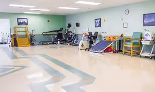 A spacious rehabilitation or physical therapy room with various exercise equipment including stationary bikes, a treadmill, parallel bars, and other therapy apparatus. The room has light-colored flooring with a geometric pattern and light green walls adorned with framed pictures and a clock.