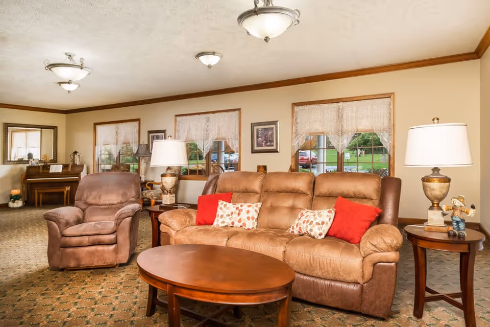 Common living area with a brown sofa and armchair, coffee table, lamps and a piano near windows.
