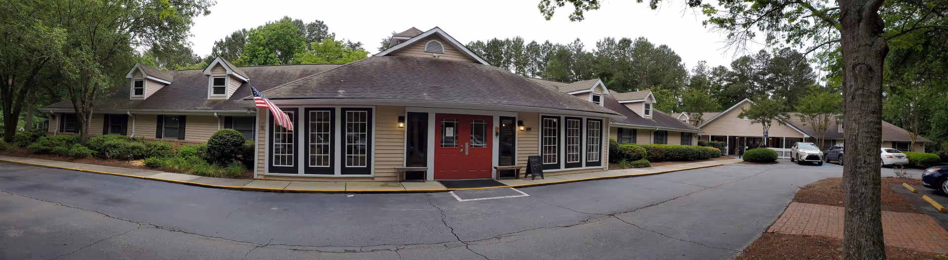 Front exterior of a single-story memory care building with a central red entrance, American flag, driveway and surrounding trees.