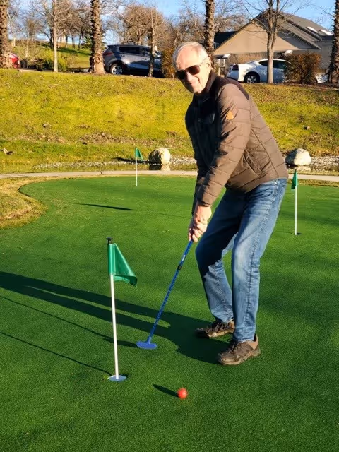 An elderly man wearing sunglasses, a brown jacket, blue jeans, and brown shoes is playing mini golf on a green putting surface. He is holding a blue putter and aiming at a red golf ball near a hole with a small green flag. The background shows a grassy hill, some trees, parked cars, and houses under a clear sky.