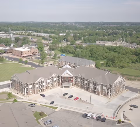 Aerial view of a large assisted living facility building with multiple floors, surrounded by parking lots and greenery. The building has a central entrance with a peaked roof and several windows. Nearby are other buildings, roads, and a green landscape extending into the distance under a cloudy sky.