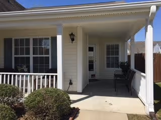 Covered front porch of a single-story residential building with columns, a railing, two chairs and shrubs.