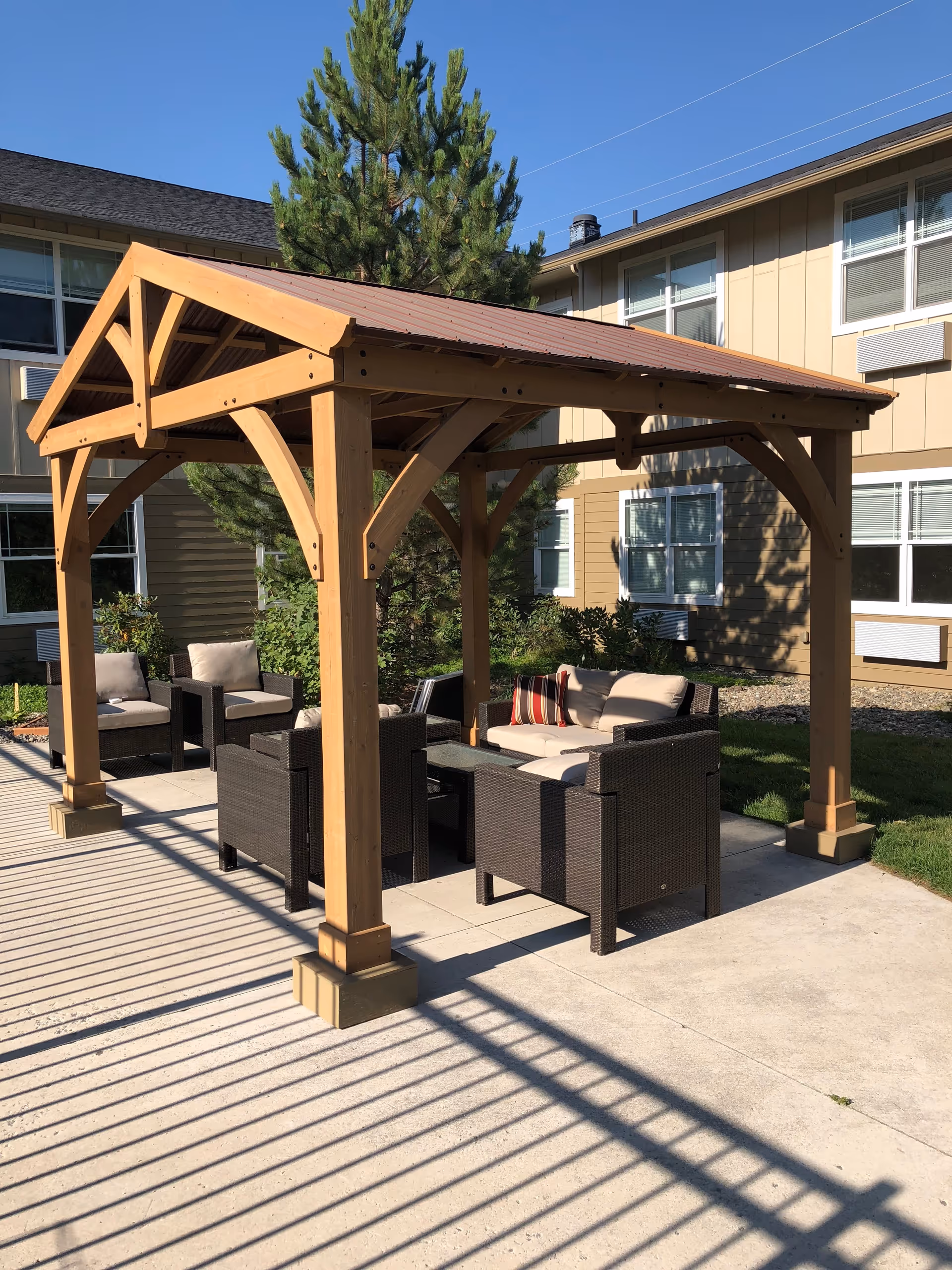 Outdoor seating area with a wooden pergola providing shade over cushioned wicker chairs and a small table, situated on a concrete patio next to a beige building with multiple windows and surrounded by greenery under a clear blue sky.