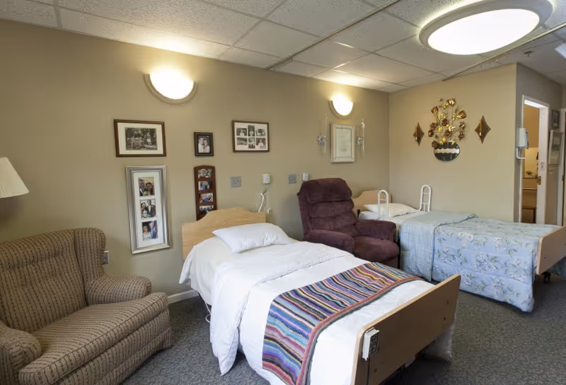 A shared senior bedroom with two hospital-style beds, an armchair and recliner, and framed photos on the wall.