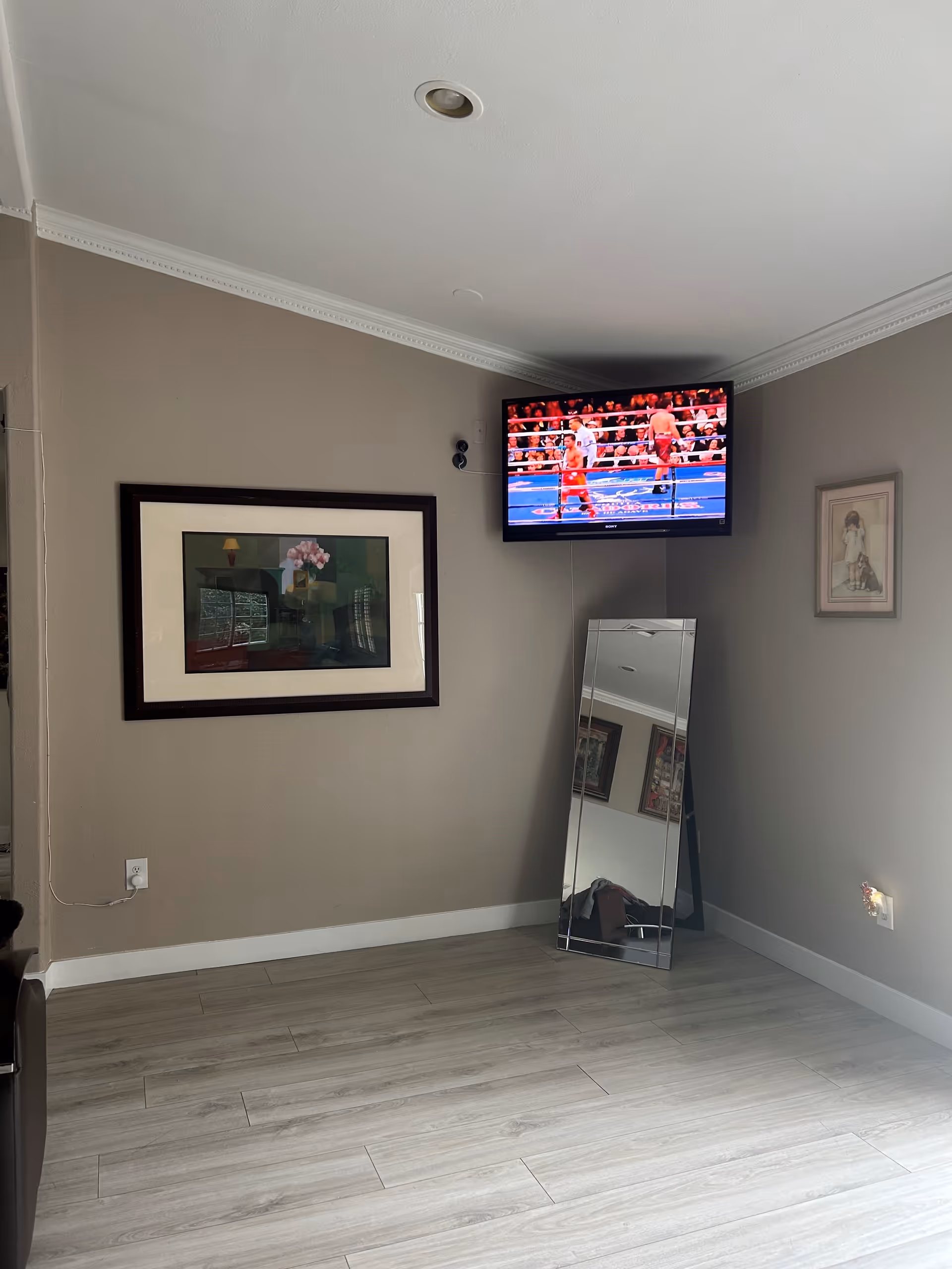 A corner of a room with light gray walls and light wood flooring. A flat-screen TV mounted on the wall shows a boxing match. Below the TV, a tall rectangular mirror leans against the wall. There are two framed pictures hanging on the walls, one larger with a floral image and one smaller with a child and dog illustration. A power outlet with a plugged-in cord is visible near the floor.