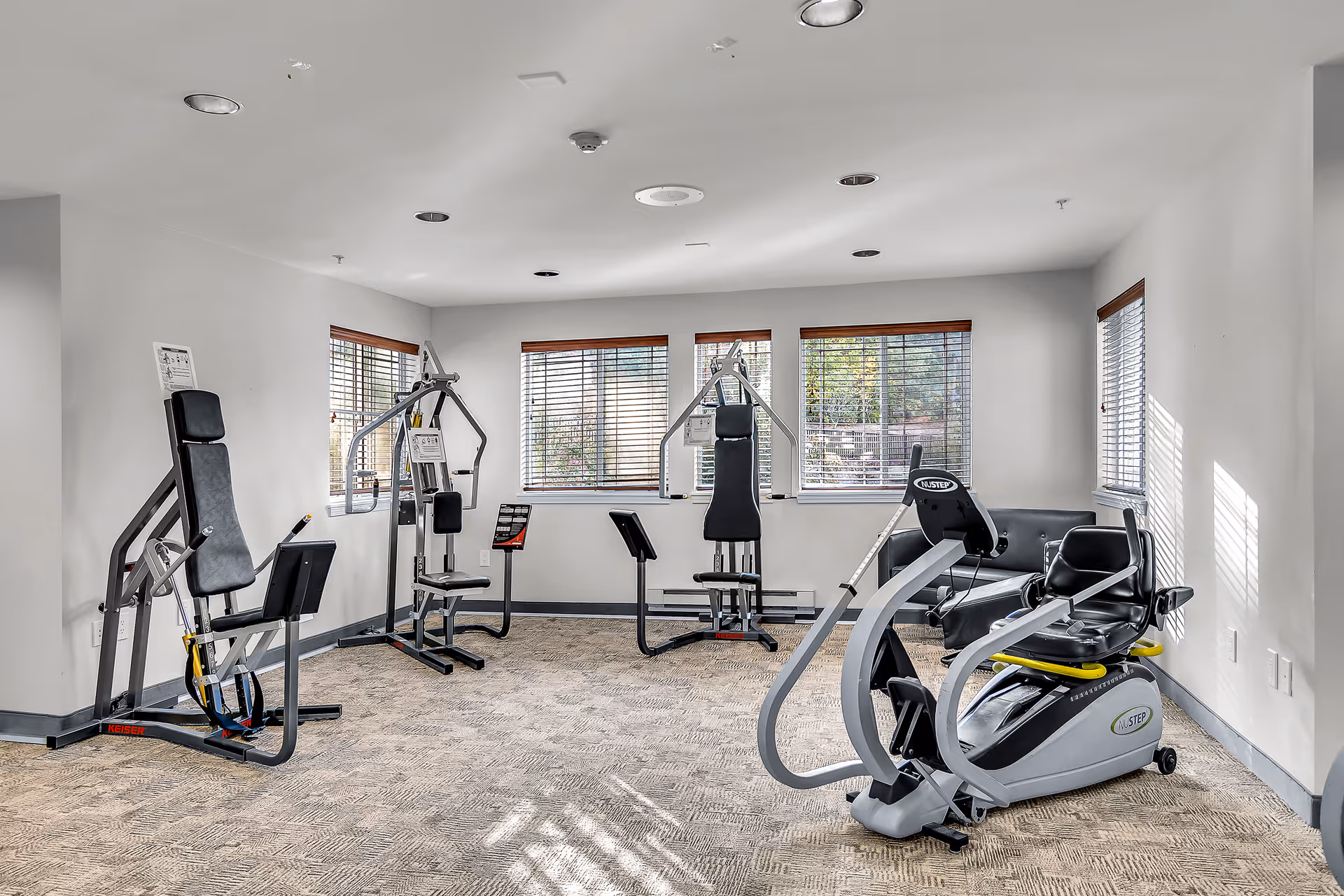A bright exercise room with several pieces of fitness equipment including weight machines and a seated elliptical machine. The room has beige carpet, white walls, and multiple windows with wooden blinds allowing natural light to enter.