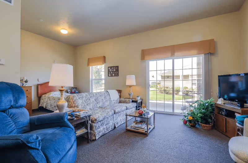 A cozy living area in an assisted living facility featuring a blue recliner chair, a floral-patterned sofa, a glass coffee table with magazines, two table lamps, a television on a wooden stand, and a sliding glass door leading to an outdoor garden area. A bed is partially visible in an adjacent room.