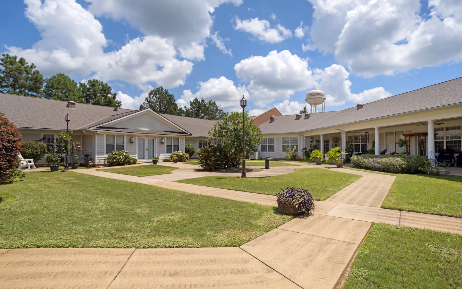 Courtyard view of a single-story senior care facility with paved walkways, lawns, shrubs and a central lamppost under a partly cloudy sky.