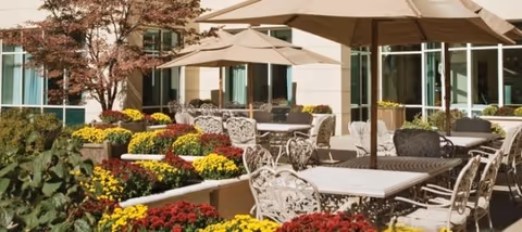 Outdoor patio area with multiple tables and chairs under large beige umbrellas, surrounded by colorful flower beds with red, yellow, and green plants, adjacent to a building with large windows.