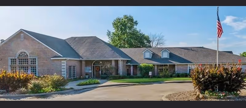 Exterior view of a single-story brick building with a sloped roof, large windows, and an American flag on a flagpole in front. The building is surrounded by landscaped greenery and a paved driveway.