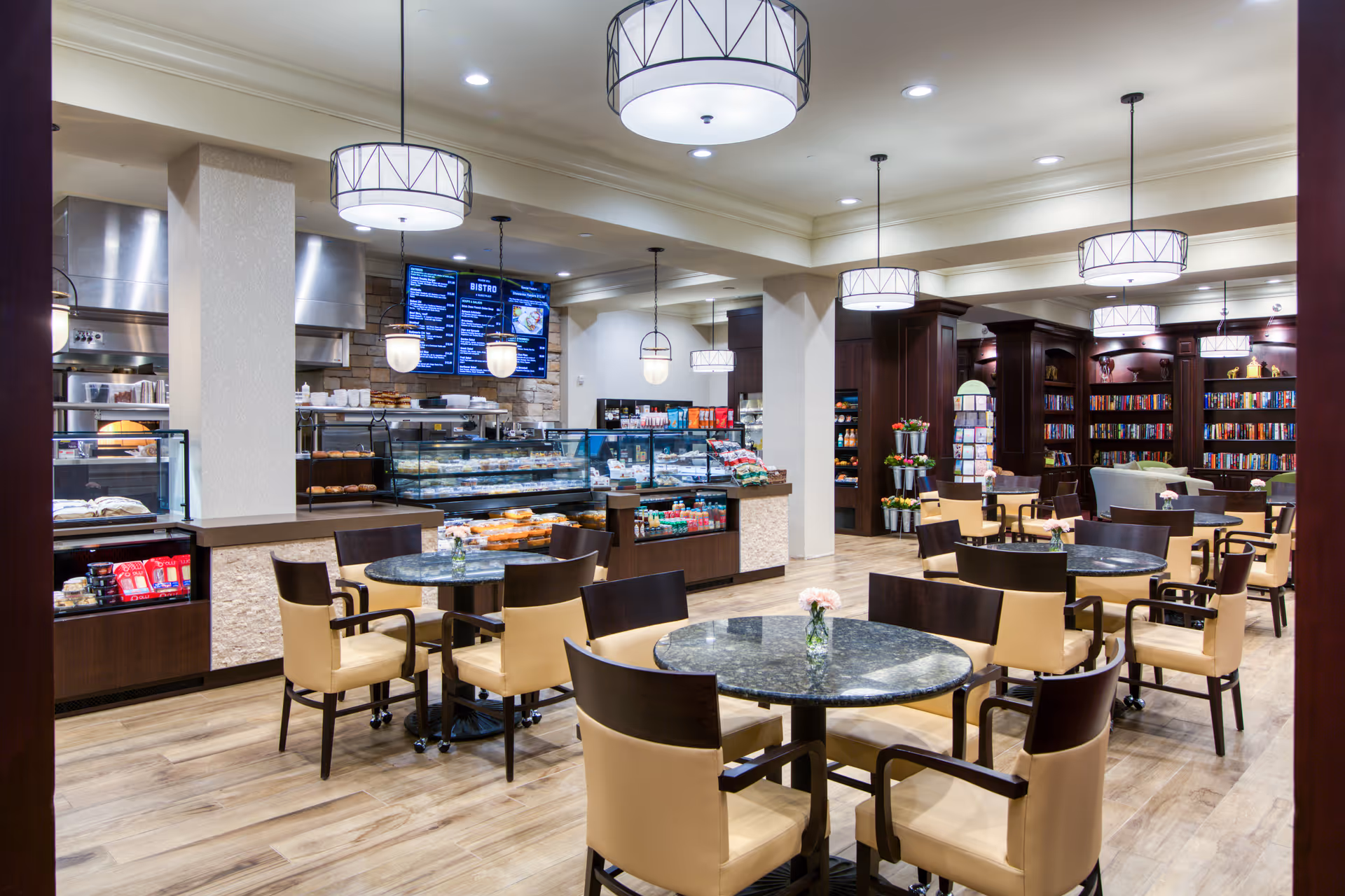 A bright and spacious dining area with round tables and beige cushioned chairs. The room features modern pendant lights hanging from the ceiling. In the background, there is a bakery counter displaying various baked goods and snacks, with a menu board above it. To the right, there is a cozy library area with bookshelves filled with books and comfortable seating.