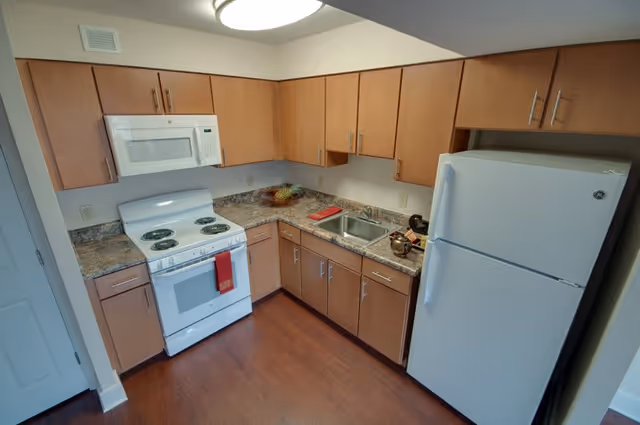A compact kitchen with wooden cabinets, a white electric stove with four burners and an oven, a white microwave above the stove, a double sink with a faucet, a white refrigerator, and a countertop with a fruit basket and some kitchen items. The floor is wooden and the walls are light-colored.