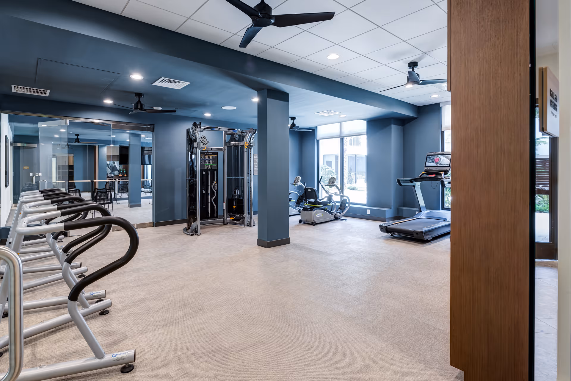 A modern fitness room with exercise equipment including treadmills, stationary bikes, and a multi-function weight machine. The room has blue walls, large windows letting in natural light, ceiling fans, and mirrors on one wall.