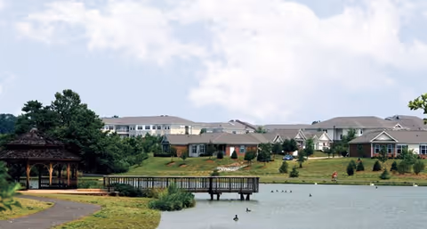 Lakeside view of Croasdaile Village showing a wooden gazebo and dock in the foreground with residential buildings and landscaping across the water.