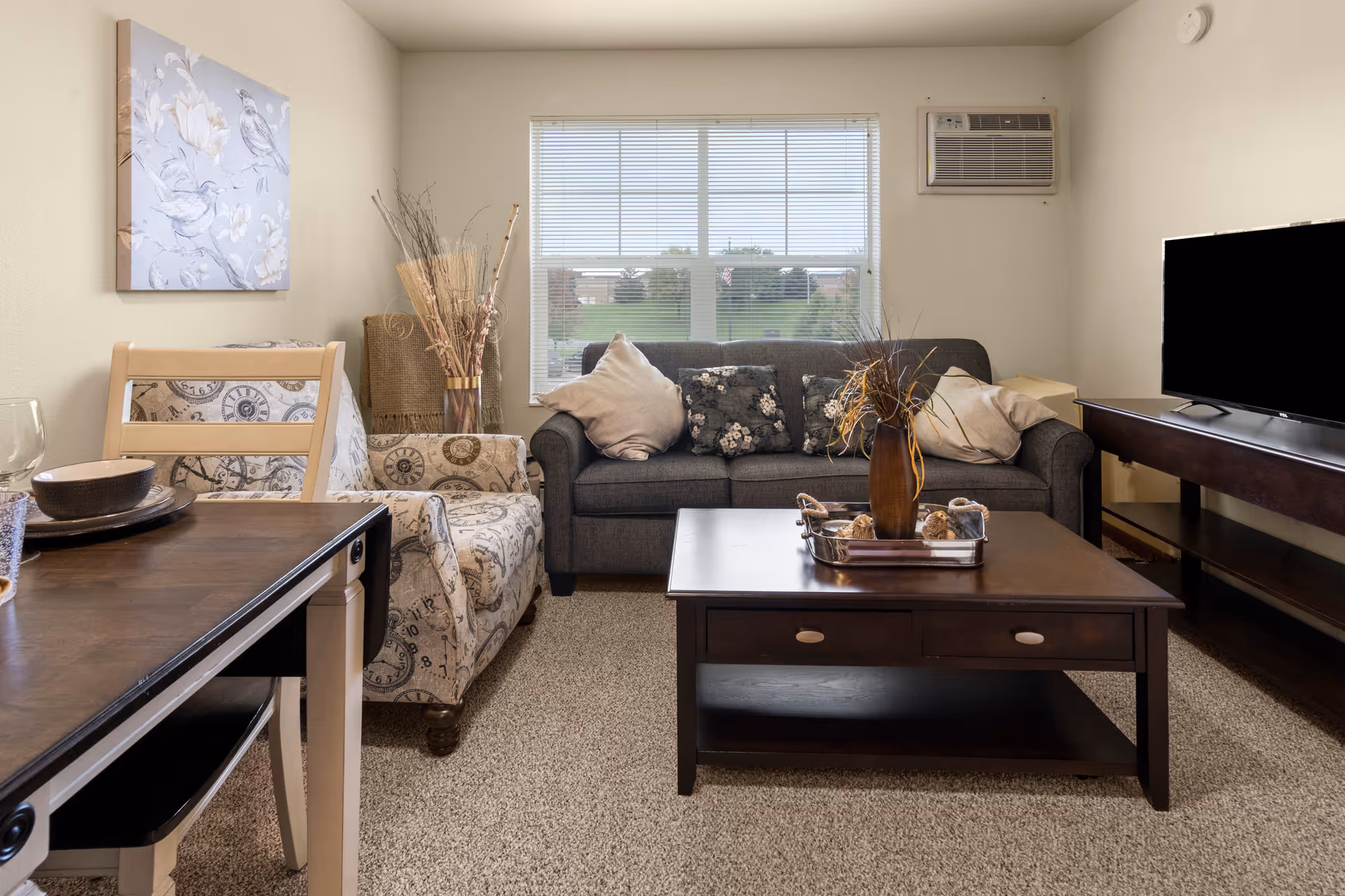 A cozy living room with a dark gray sofa adorned with decorative pillows, a patterned armchair, a wooden coffee table with a vase and decorative items, a TV on a wooden stand, a dining table with chairs, and a window with blinds letting in natural light.