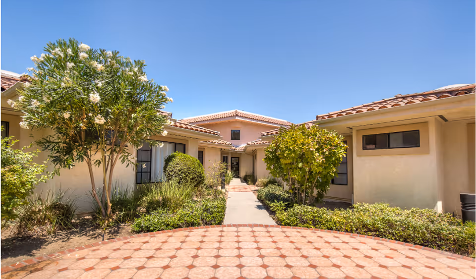 Courtyard entrance to a single-story stucco building with tiled roofs, a paved walkway and landscaping under a clear blue sky.