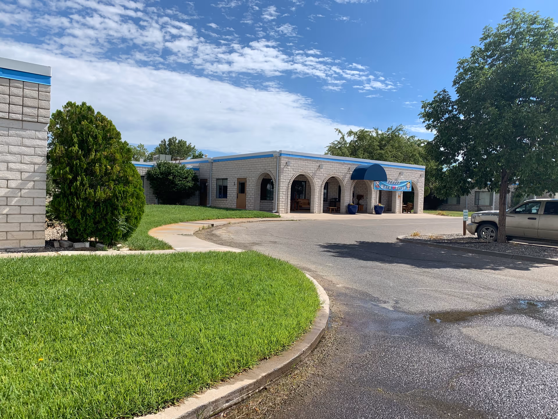 Single-story brick building with arched covered entrance and blue awning, a curved driveway, parked vehicle, and green lawn under a partly cloudy sky.