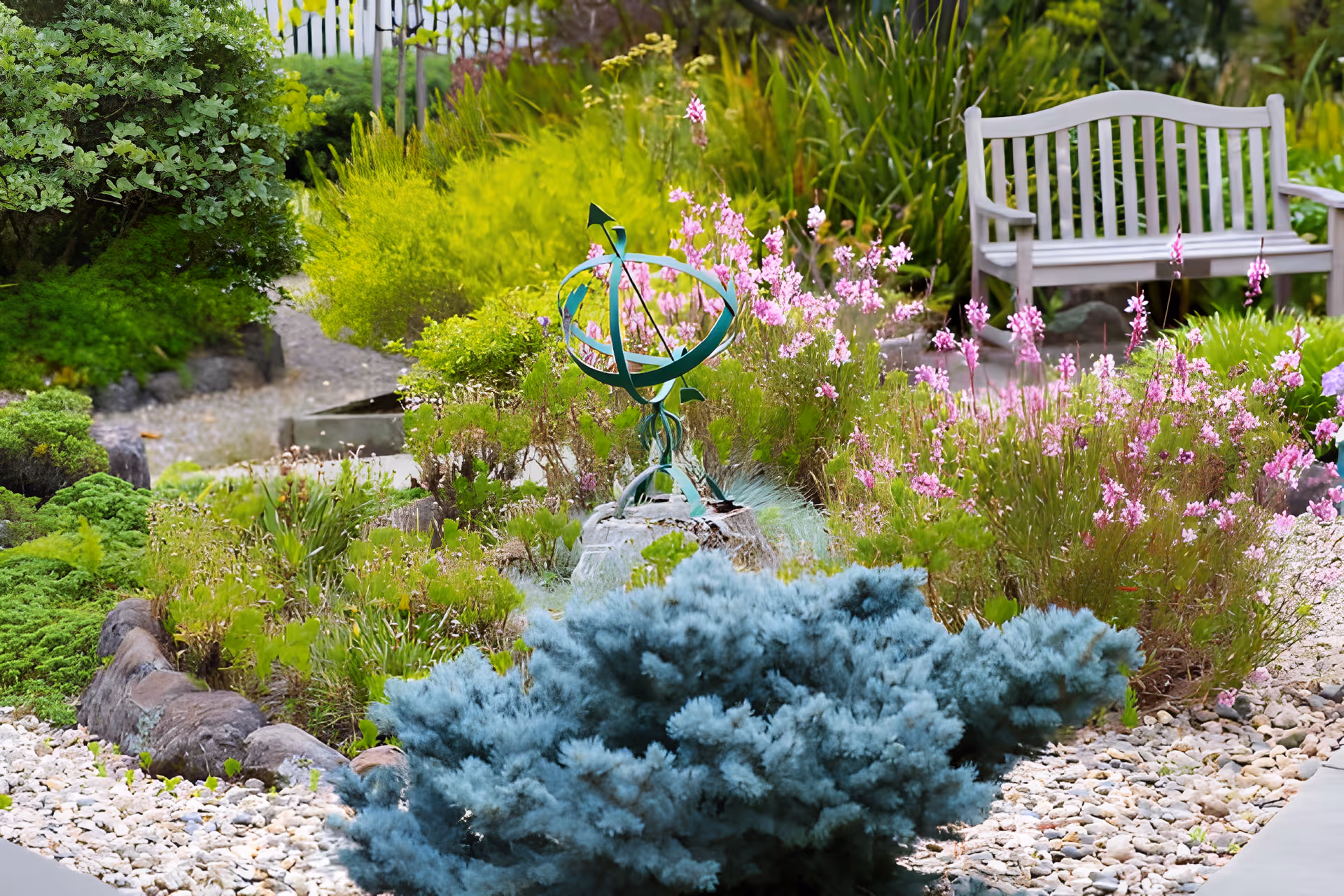 A landscaped garden with flowering plants, a decorative sundial sculpture, gravel paths, and a wooden bench.