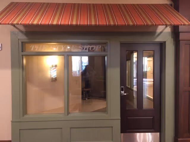 Entrance to a barbershop with a striped red and orange awning above. The door is dark brown with glass panels, and there is a window next to it with a lit barber pole inside. The sign above the window reads 'THE STYLE SHOP'.