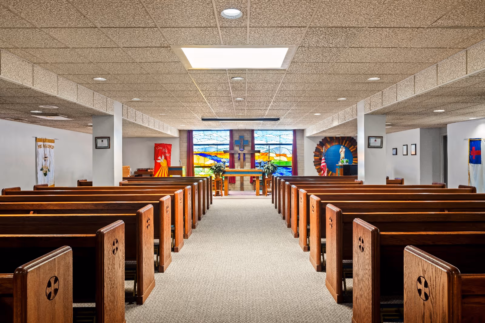 Interior view of a chapel with wooden pews arranged in rows facing an altar. The altar is positioned in front of a large stained glass window featuring colorful designs and a cross. The ceiling has recessed lighting and a skylight, and religious banners hang on the walls.