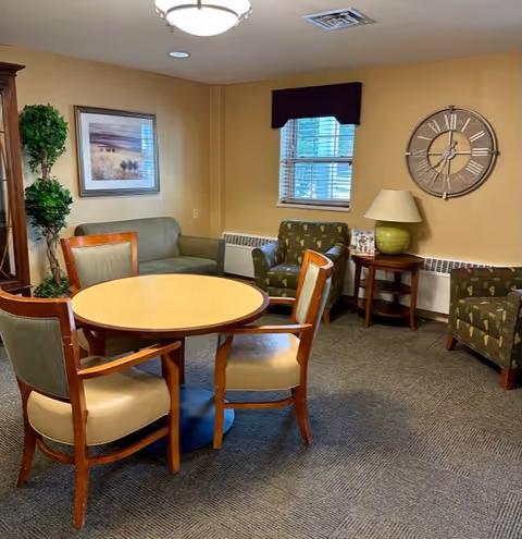 A cozy sitting area in a senior living facility with a round wooden table surrounded by four chairs. There is a green loveseat, two patterned armchairs, a side table with a lamp, a large wall clock, a framed landscape picture, and a potted plant near the corner. A window with blinds and a dark valance lets in natural light.