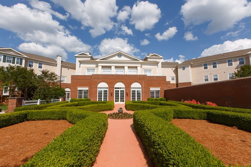 A large senior living facility building with a well-maintained garden featuring neatly trimmed hedges and a brick pathway leading to the entrance. The building has a combination of red brick and light-colored siding with multiple windows and a blue sky with scattered clouds above.