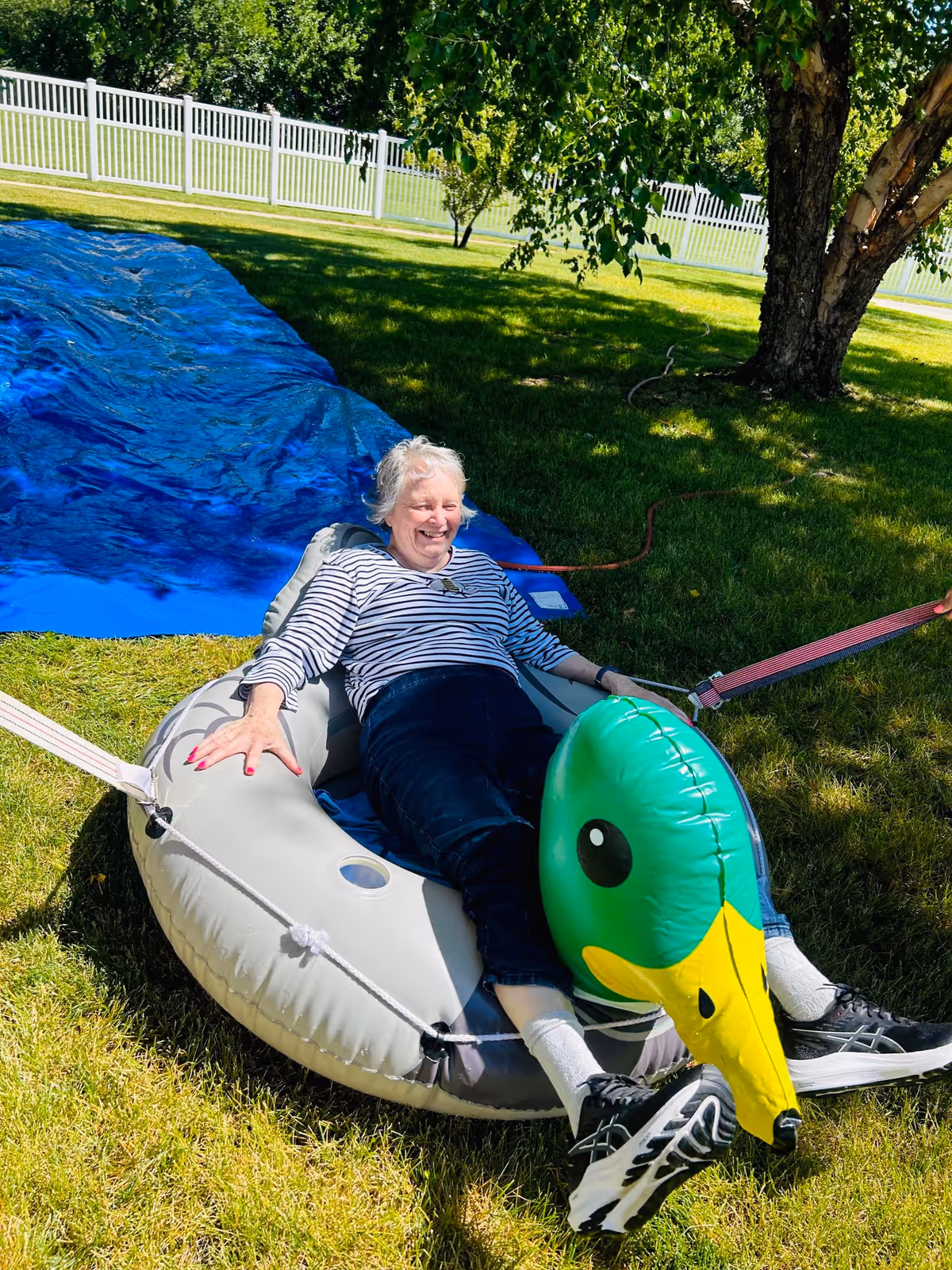 An elderly woman with short white hair, wearing a striped long-sleeve shirt and dark pants, is sitting and smiling on an inflatable raft with a green duck head at the front. The raft is on grass near a blue tarp slide, with a white fence and trees in the background on a sunny day.