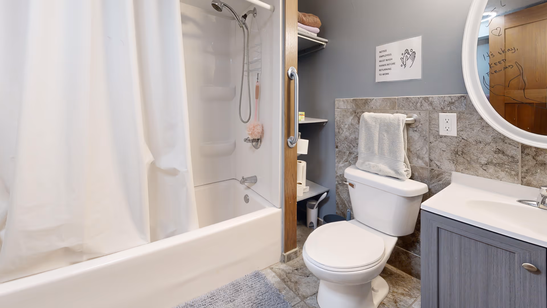 A clean bathroom featuring a white bathtub with a showerhead and a white shower curtain. Next to the bathtub is a white toilet with a folded towel hanging on a grab bar above it. The wall behind the toilet has gray tiles and a sign reminding to wash hands. A gray vanity with a white sink and a round mirror above it is visible on the right side. The floor is tiled and a gray bath mat is placed in front of the tub.