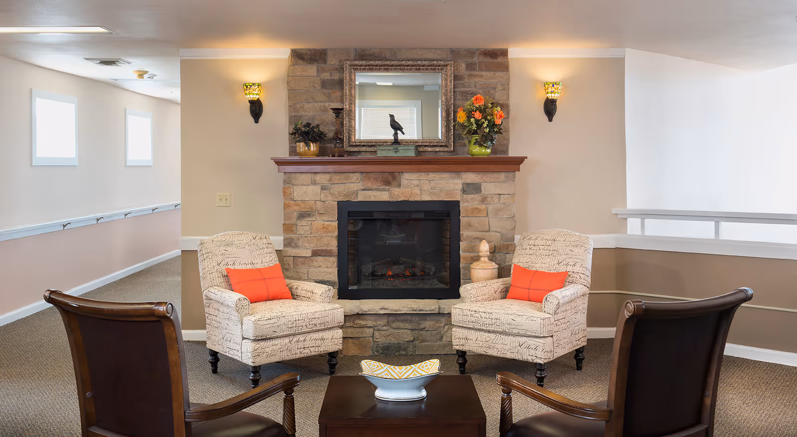 A cozy sitting area with a stone fireplace in the center, flanked by two upholstered armchairs with orange cushions. Two dark wooden chairs face the armchairs across a small wooden table with a decorative bowl. The walls are painted beige and white, with two wall sconces providing warm lighting.
