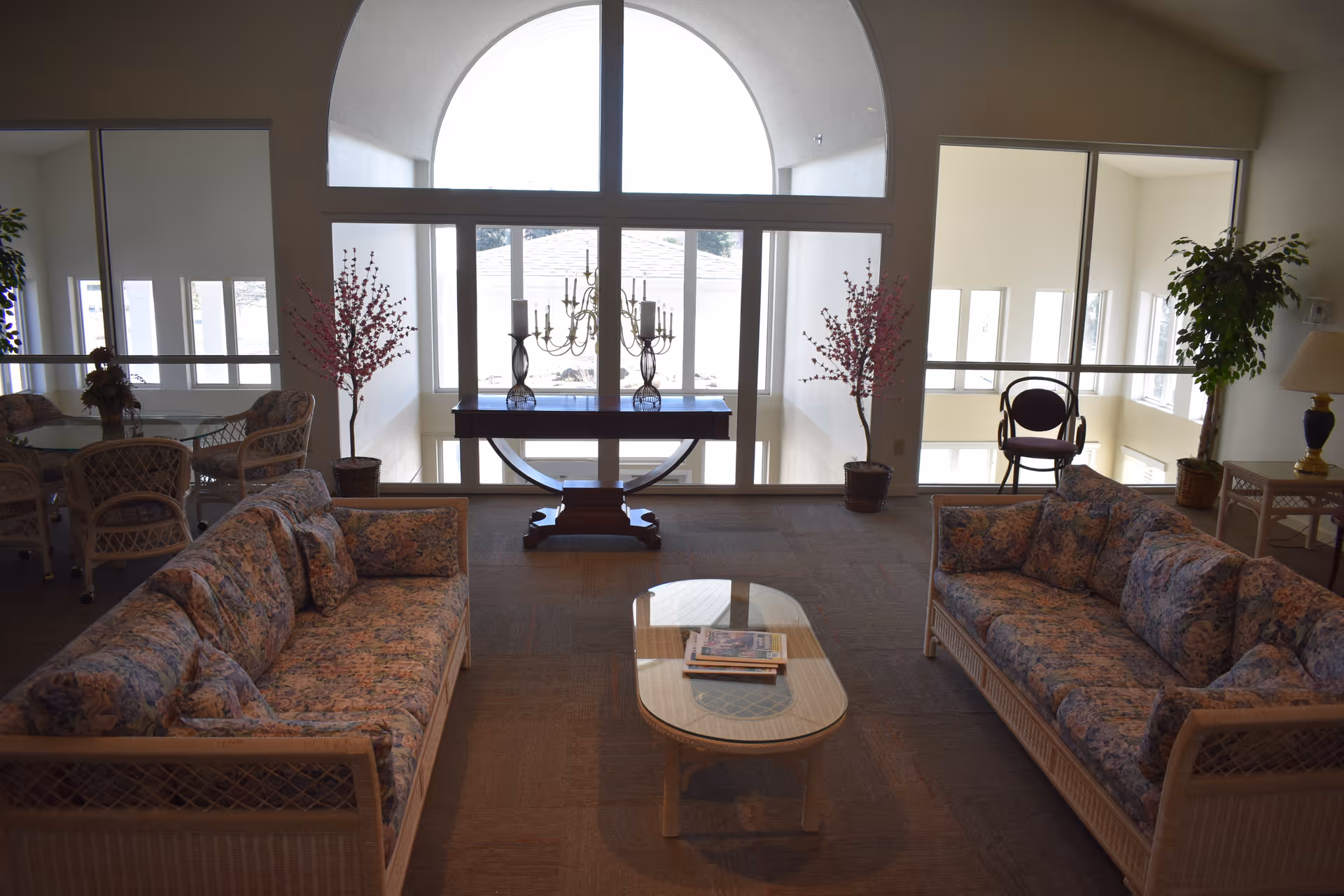 Sunlit communal living room with two floral-patterned sofas facing a glass coffee table beneath a large arched window and decorative table with candelabras.