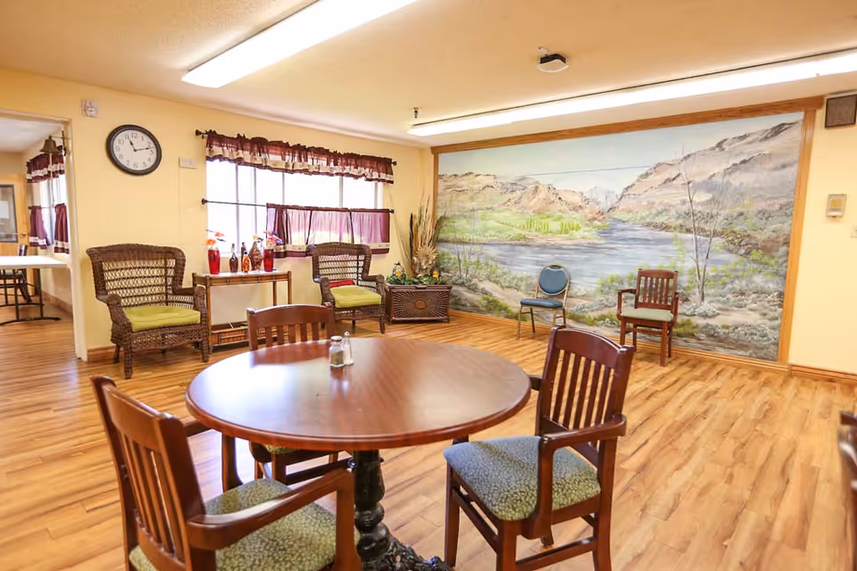 Bright common dining area with a round wooden table and chairs, wicker seating, and a large landscape mural on the wall.