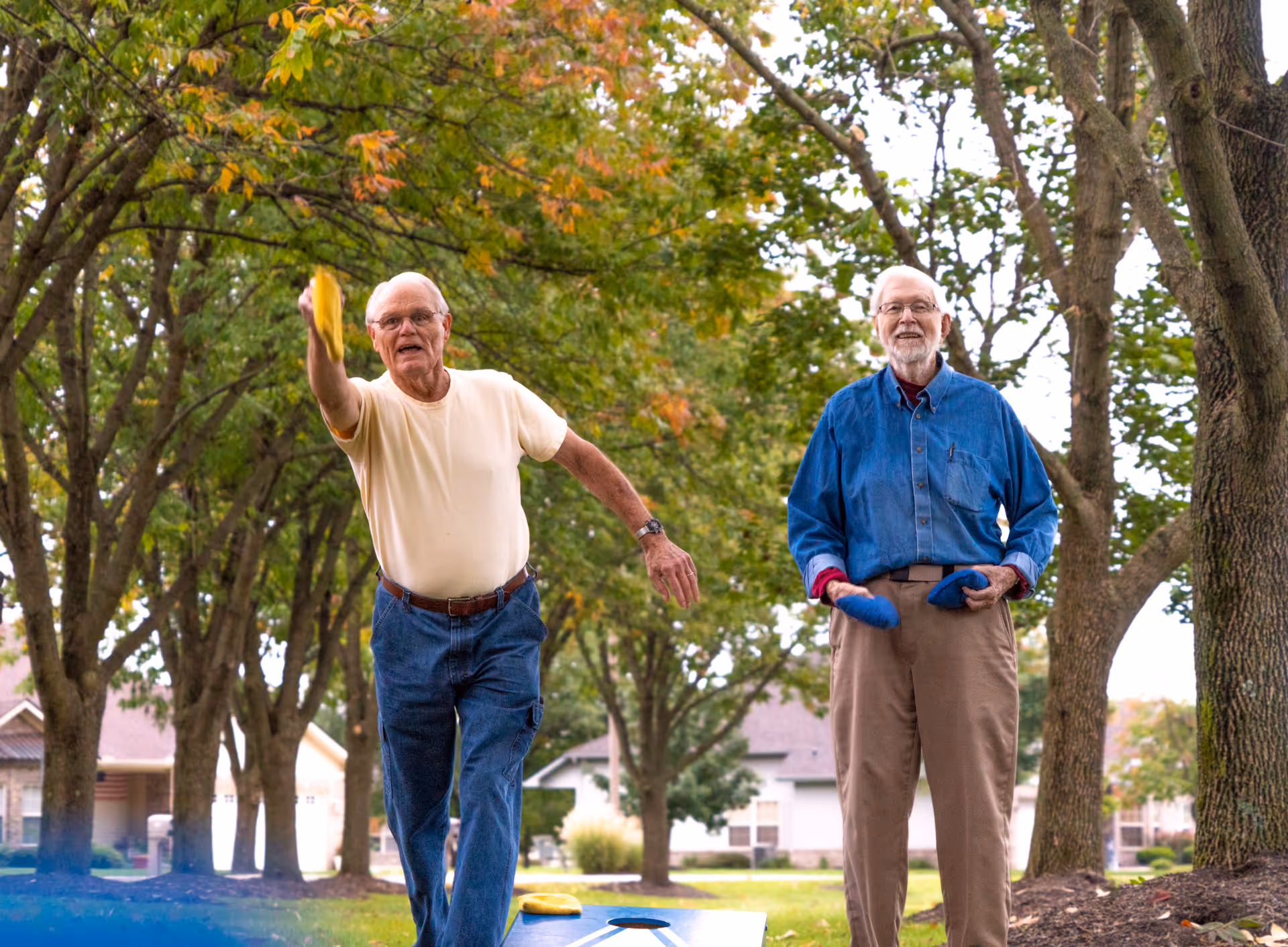 Two elderly men playing cornhole outdoors in a tree-lined area with houses in the background. One man is in mid-throw wearing a light yellow shirt and blue jeans, while the other man stands holding bean bags, dressed in a blue shirt and beige pants.
