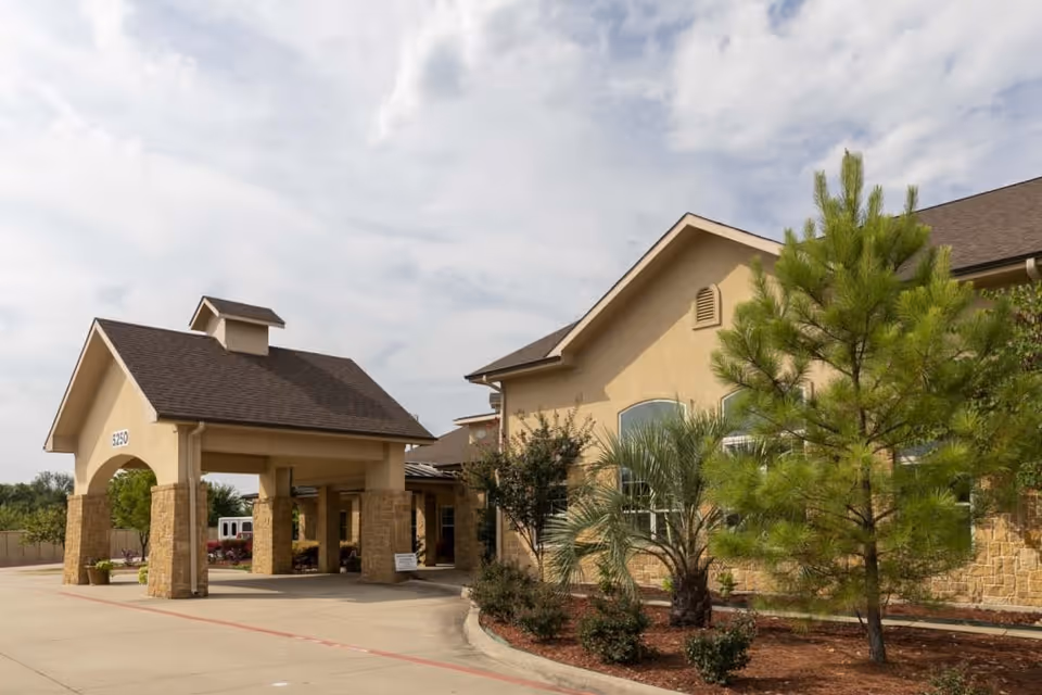 Exterior view of a senior living facility with a covered entrance supported by stone pillars, beige walls, and a dark shingled roof. There are landscaped plants and trees along the driveway under a partly cloudy sky.