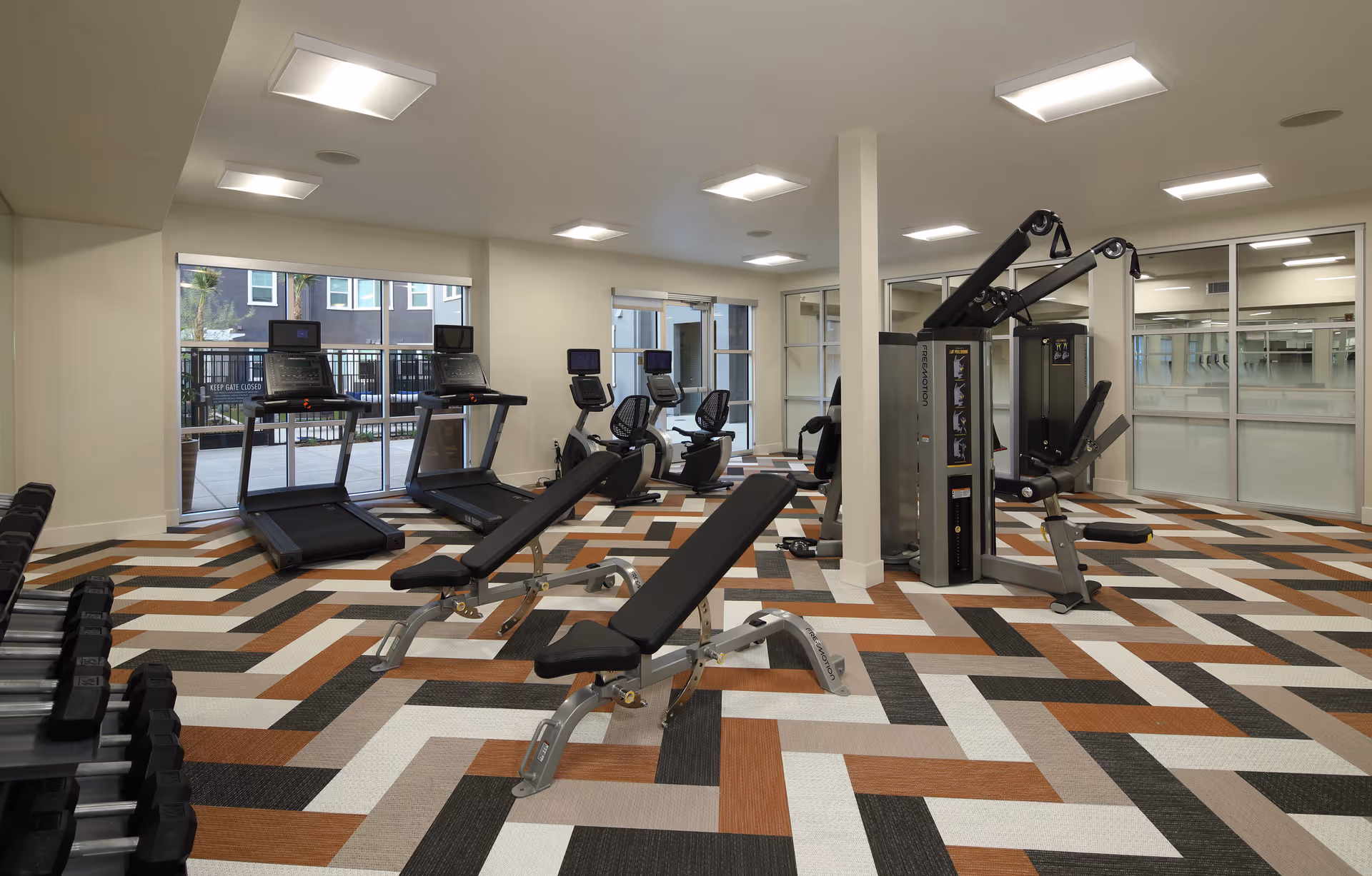 Well-lit fitness room with treadmills, weight benches, strength machines, and a patterned floor.