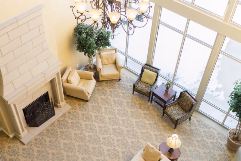 Bright senior living lounge with fireplace, chandelier, and expansive windows.