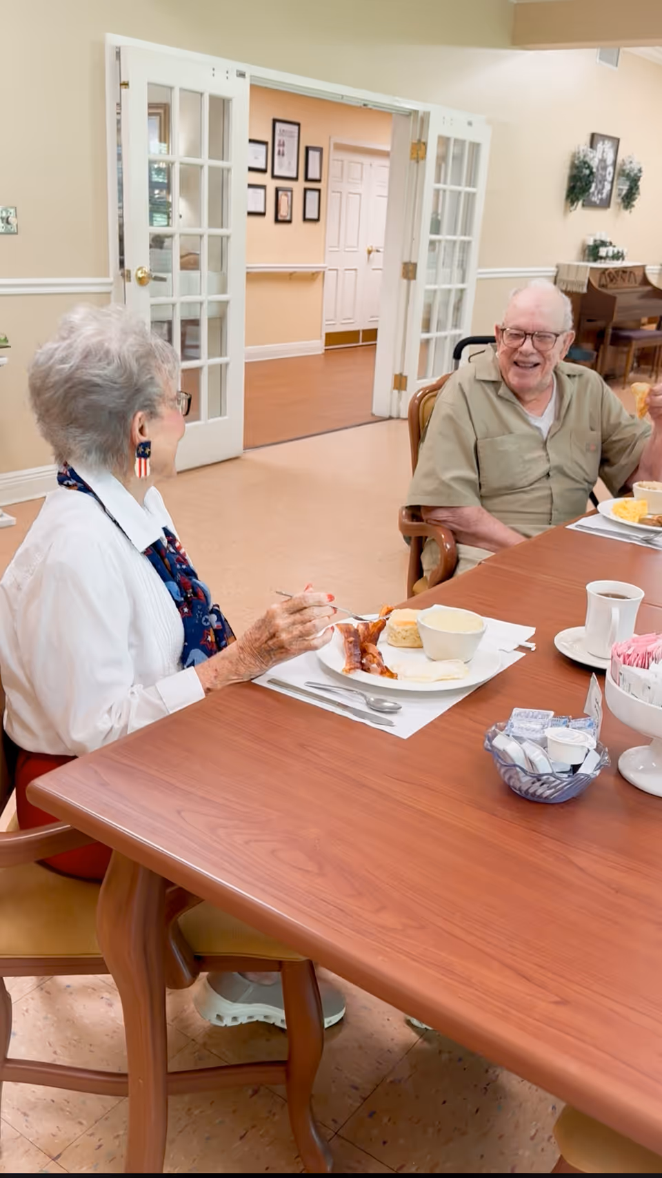 Two elderly residents smiling and eating together at a dining table inside an assisted living facility.