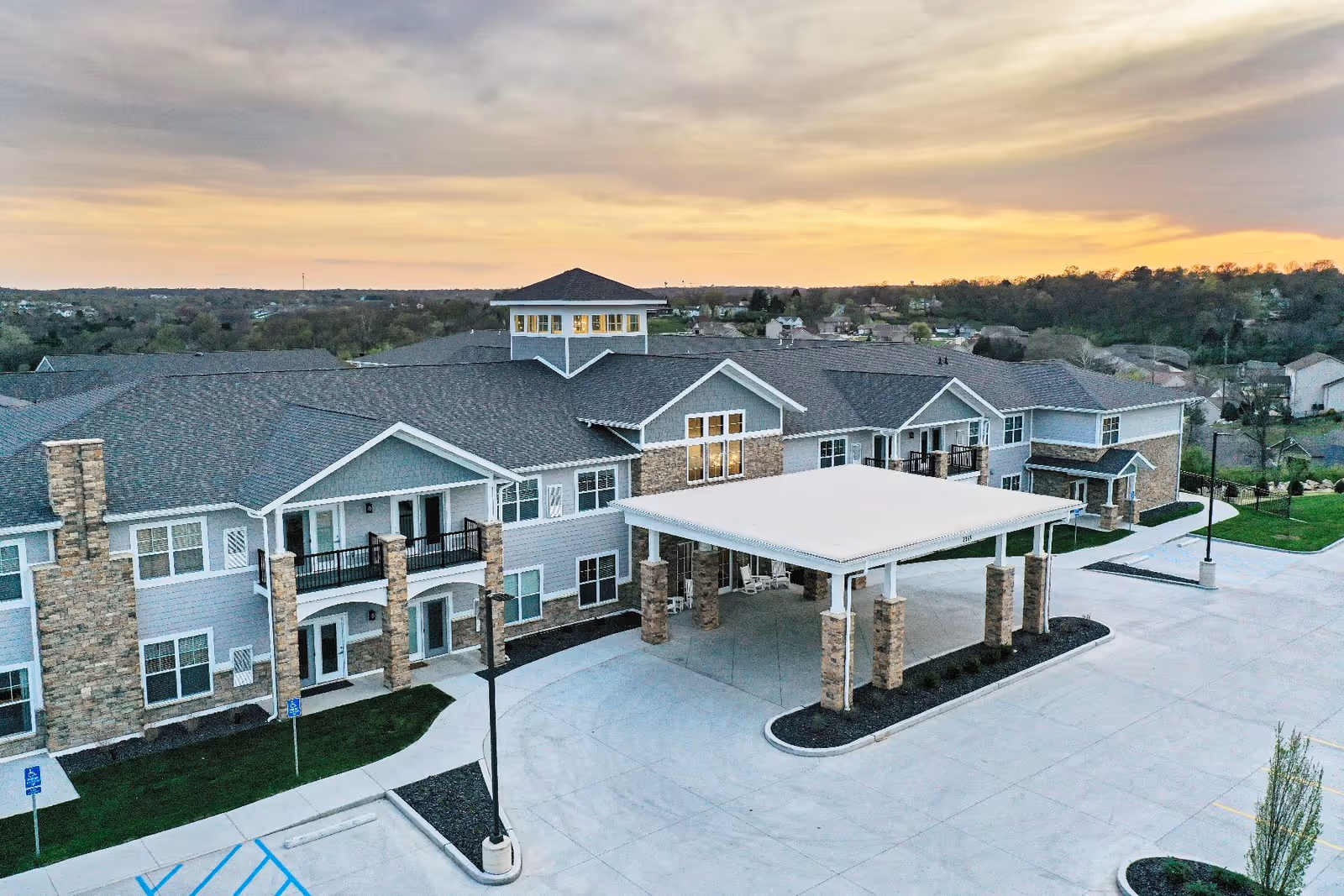 Aerial view of a two-story senior living building with a covered porte-cochere entrance, parking lot, and sunset sky.