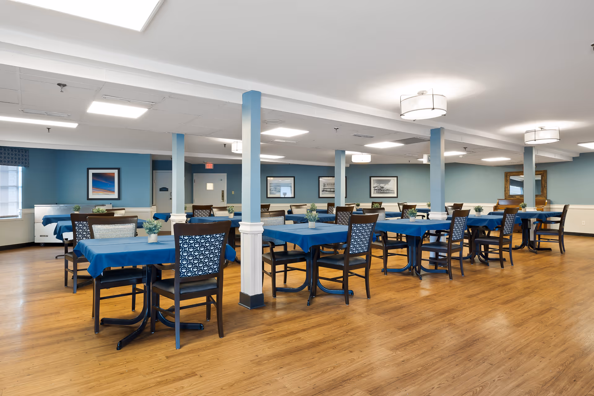 A spacious dining room with multiple tables covered in blue tablecloths, each surrounded by chairs. The room has light blue walls, wooden flooring, ceiling lights, and framed pictures on the walls. Small potted plants are placed on each table.